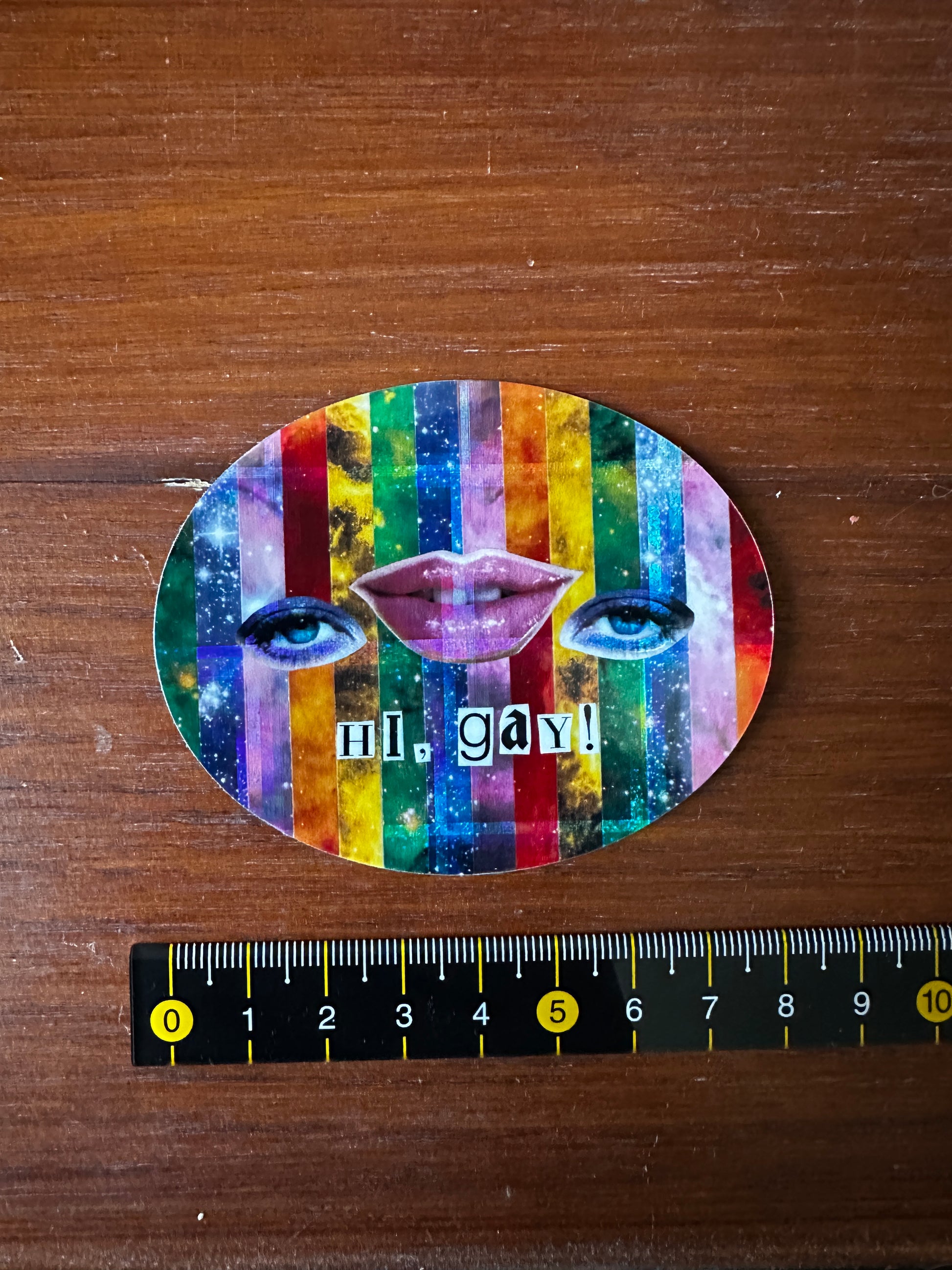 Colorful face-shaped sticker with 'hi, gay!' text on a wooden surface with a ruler for scale.