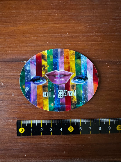 Colorful face-shaped sticker with 'hi, gay!' text on a wooden surface with a ruler for scale.